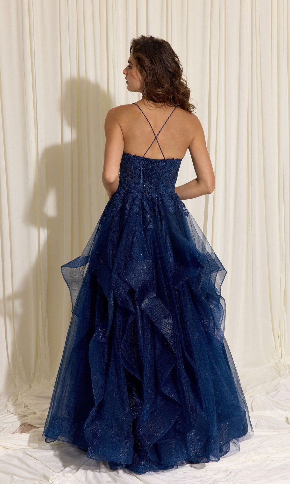 A woman poses with her back to the camera, wearing the Eureka EK167 long prom dress—a dark blue, floor-length glitter tulle gown with thin crisscross straps and a layered A-line skirt—set against a light curtain backdrop.
